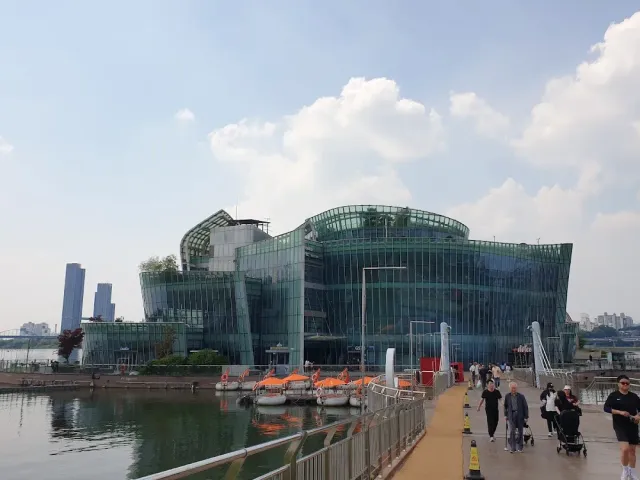 A large glass structure on the water, a promenade with walkers and orange boats