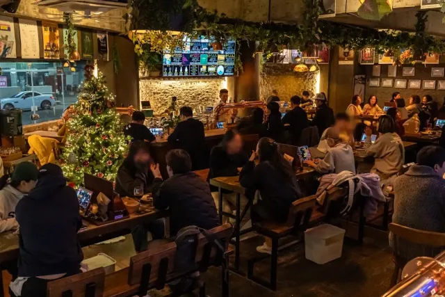 A busy pub with full wooden tables, a bar with taps and a decorated fir tree
