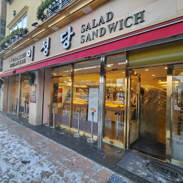 A bakery front with a burgundy sign and lit display windows, a snowy sidewalk