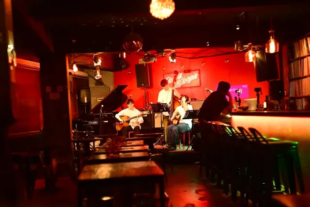 A club in red light with musicians on a small stage, a grand piano and a bar with stools