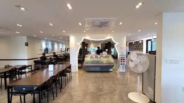 A bright hall with long wooden tables, a tiled floor and a lit refrigerated counter at the back