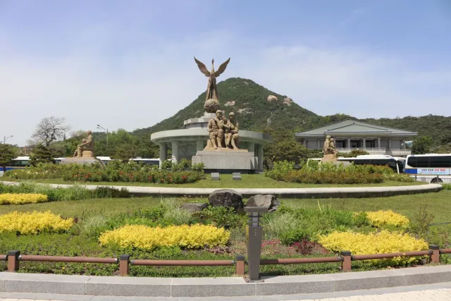 A bronze memorial with seated figures and a winged statue, surrounded by flower beds
