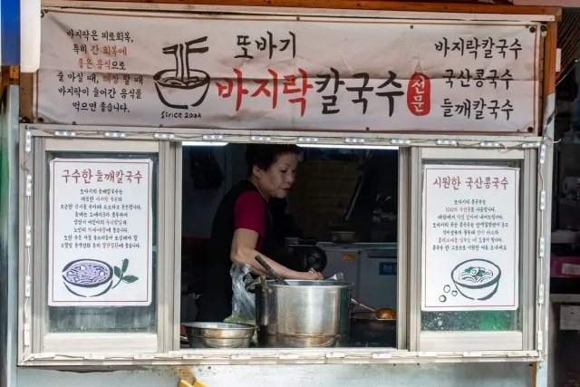 A woman cooking in a large pot at a food stall with menu posters on both sides