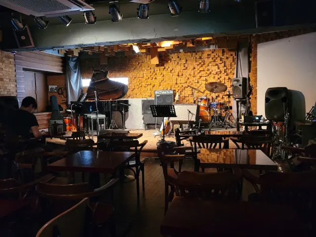 A small stage with a grand piano and a drum kit, and empty wooden chairs facing it
