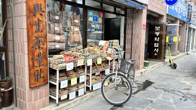 A shop front with crates of dried medicinal herbs, a carved sign and a bicycle outside