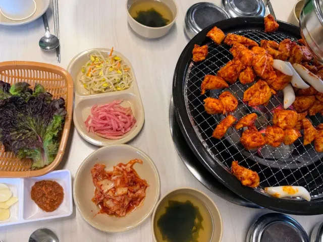 Chicken pieces in red sauce grilling on a charcoal grate, beside lettuce, kimchi and side dishes