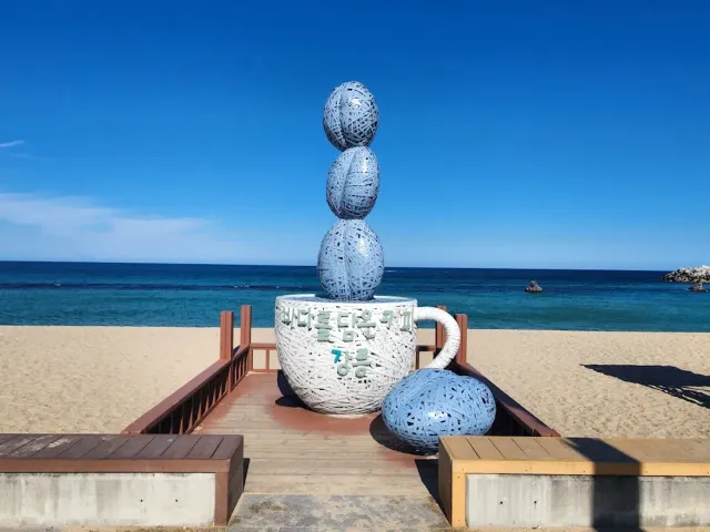 A giant coffee cup sculpture with balls stacked above it, on a sandy shore facing the sea