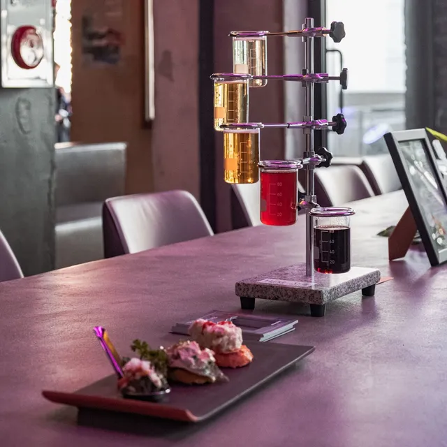 A beer sampler in glass beakers on a lab stand, beside a plate of small appetizers