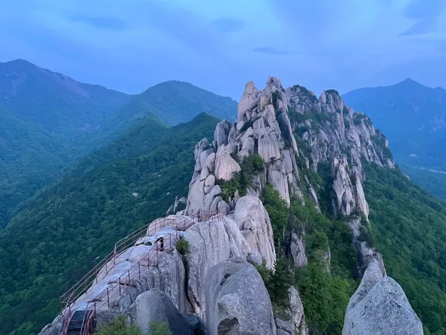A jagged granite ridge with steps and a red railing, green mountains all around