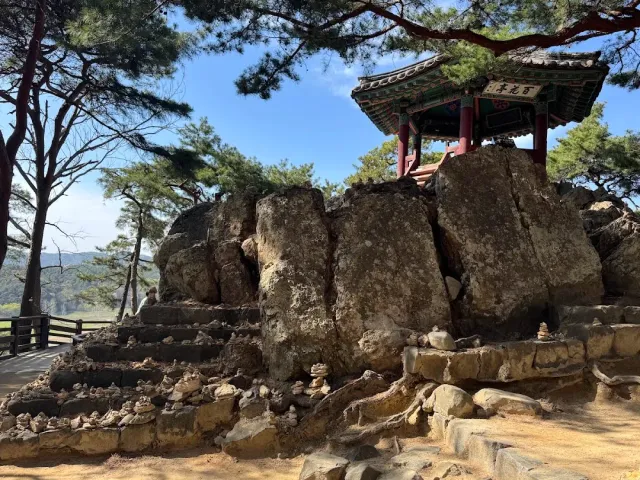 A colorful wooden pavilion on a large rock mass, pine trees and small stone cairns