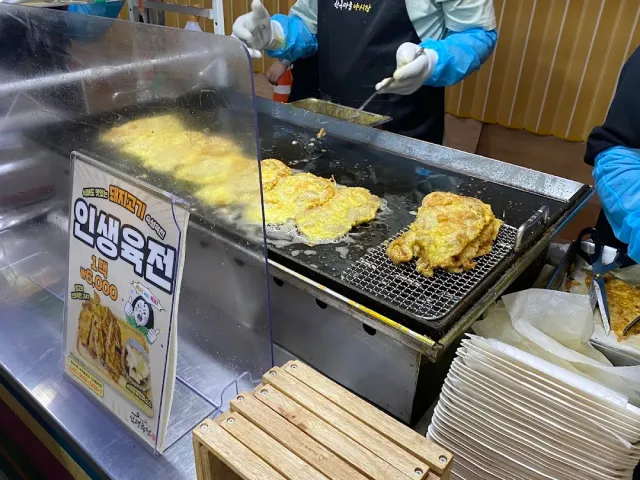 Egg battered fritters frying on a large griddle and a cook in blue gloves