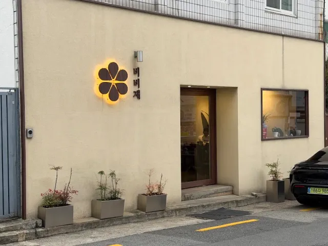 A cream coloured front with a lit flower shaped sign, a wooden door and planters along the pavement