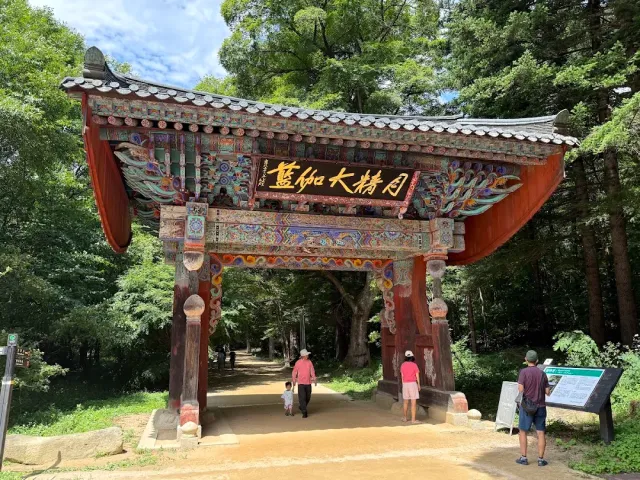 A vividly painted wooden gate with a gilded sign, and a dirt path leading into a green forest