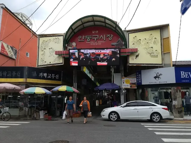 A market entrance arch with a video screen, striped umbrellas and two women crossing a road
