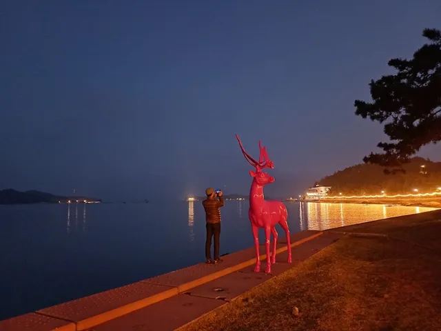 A pink deer statue on a promenade by a calm bay, with the shore lights at night