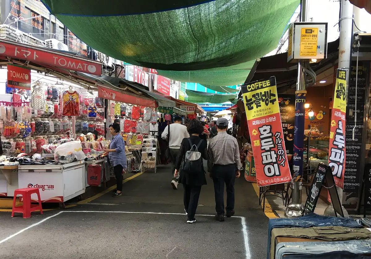 A market alley under green shade netting, with packed stalls and colourful signs on both sides