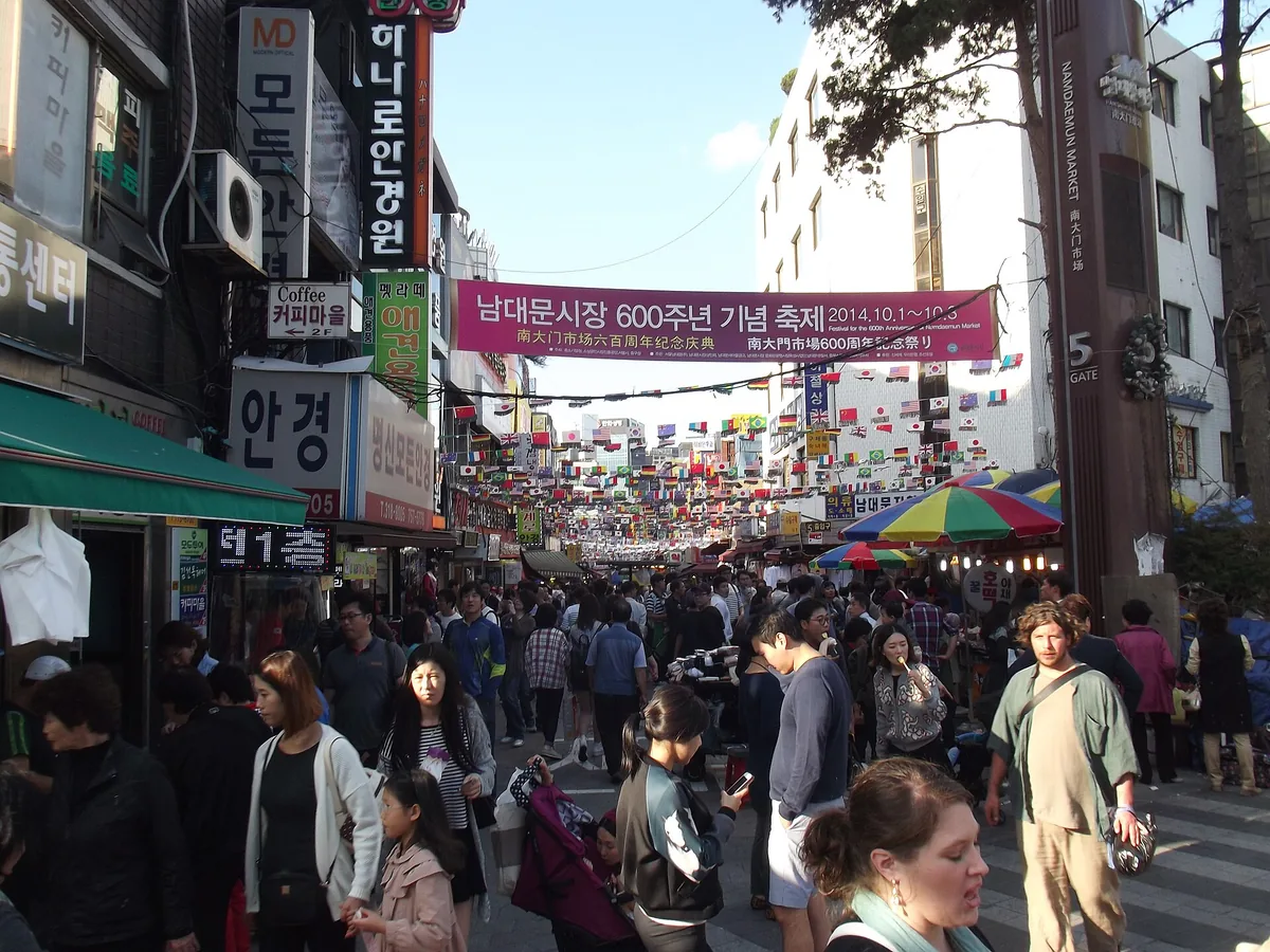 A market street packed with shoppers between sign covered storefronts, with a banner strung overhead