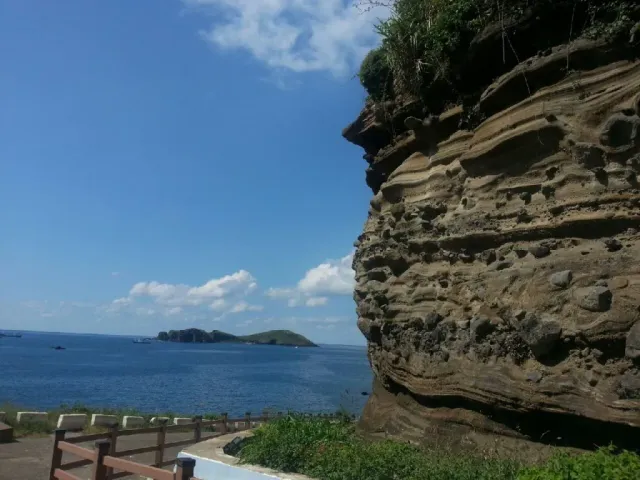A cliff of horizontal stone layers facing a blue sea with a small island on the horizon