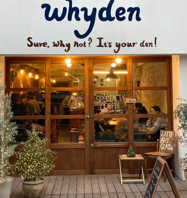 A front with a white sign, wooden windows, people sitting inside and trees lit with bulbs