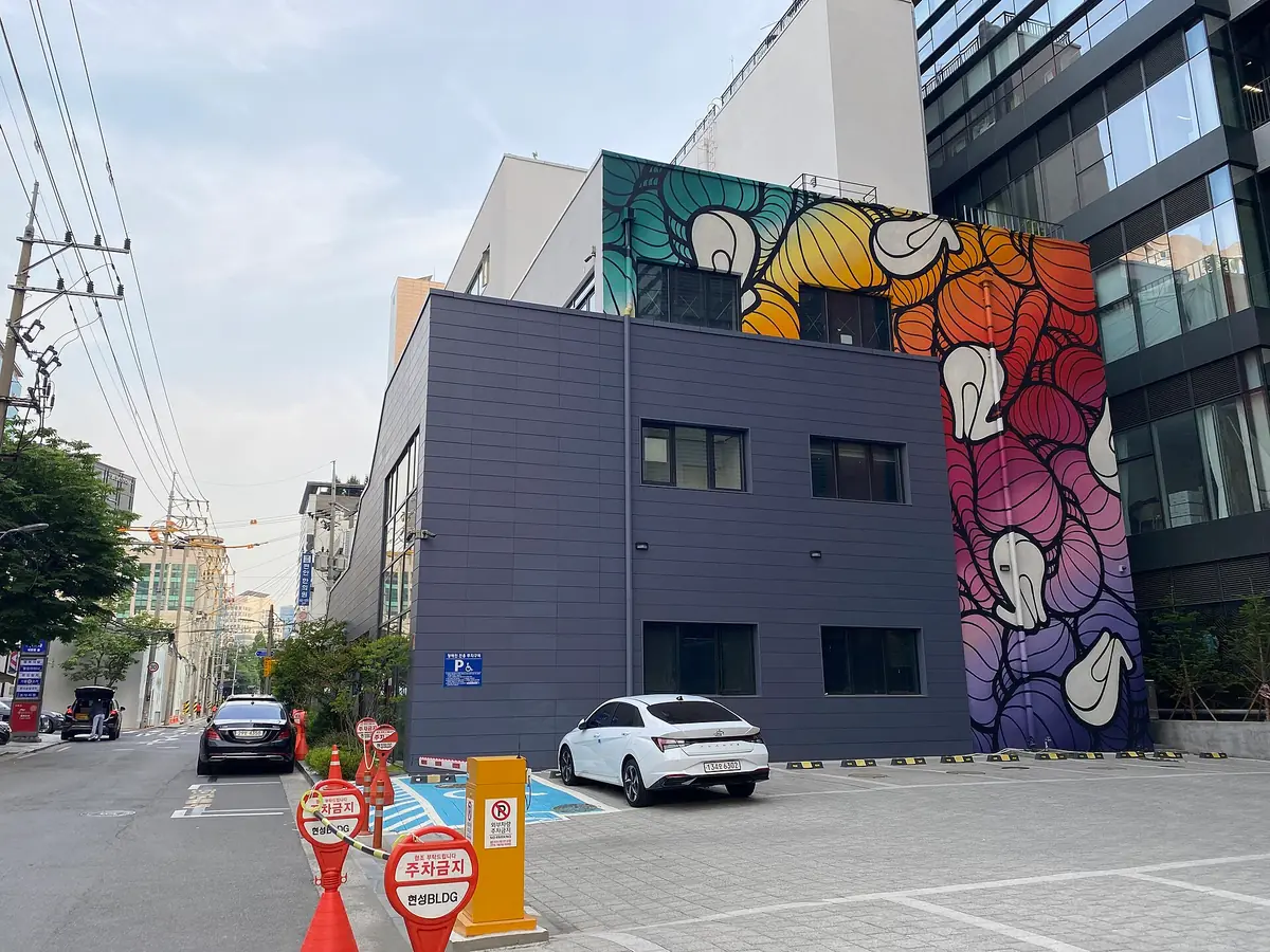 A colourful mural on a grey industrial building at a street corner, with cars parked in front