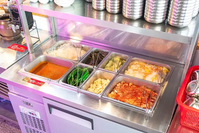 A refrigerated counter with metal trays of side dishes, and stacked metal bowls above them