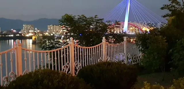 A lit cable stayed bridge over a river at evening, an ornate white fence in the foreground