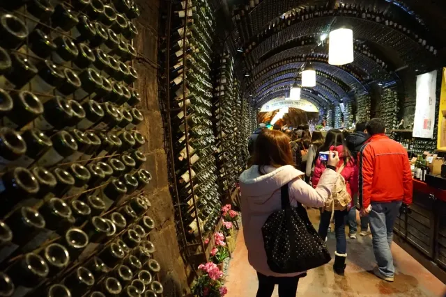 An arched tunnel with walls lined in bottles, visitors walking through it