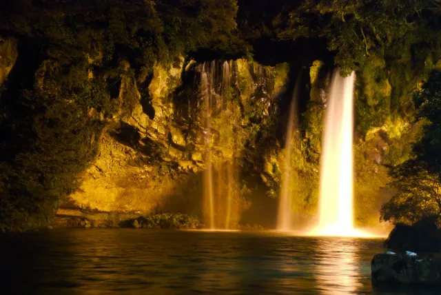 A wide waterfall lit in gold falling into a dark pool at night