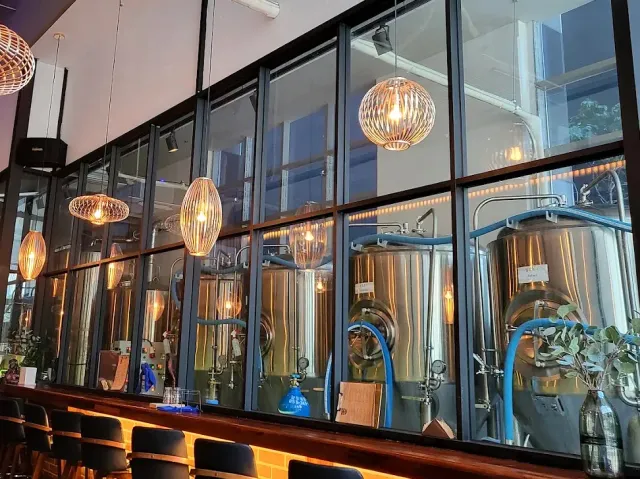 A counter with stools facing a glass wall, with stainless steel tanks behind it and woven lamps