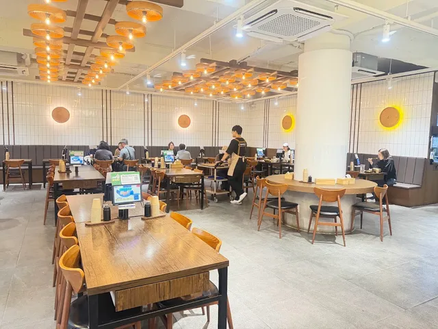 A bright restaurant hall with long wooden tables, orange chairs and hanging straw lamps