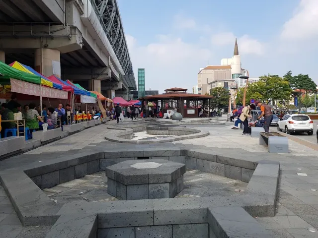 A paved plaza with sunken stone basins beneath an elevated railway, and colorful stalls to the side