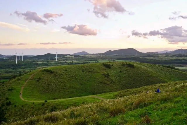A grass covered volcanic crater with walking paths, wind turbines and sunset clouds
