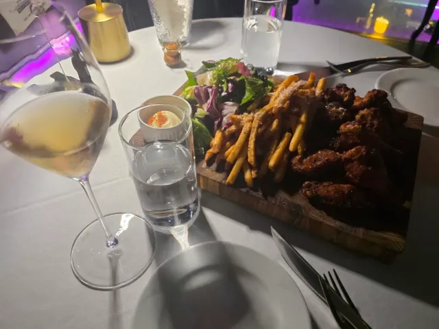 A wooden board with fried chicken and fries beside a wine glass on a white table