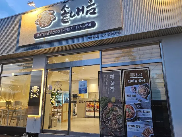 A lit front in the evening with a sign, glass windows and a banner showing photos of dishes