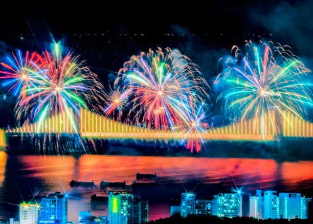 Colorful fireworks bursting above a lit bridge and their reflection in the water below