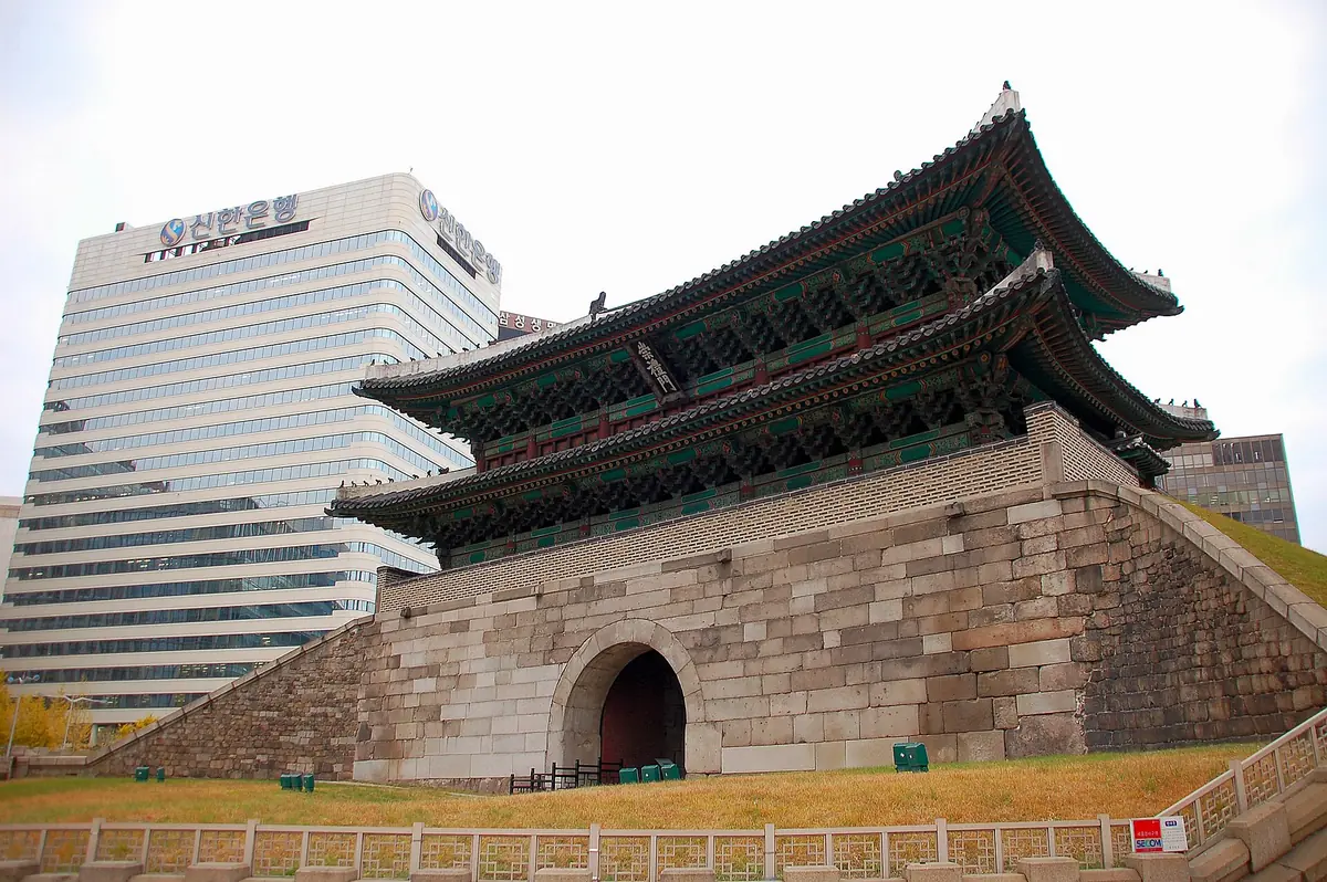 An ancient gate with a wooden pavilion on a massive stone base, and a modern office tower behind