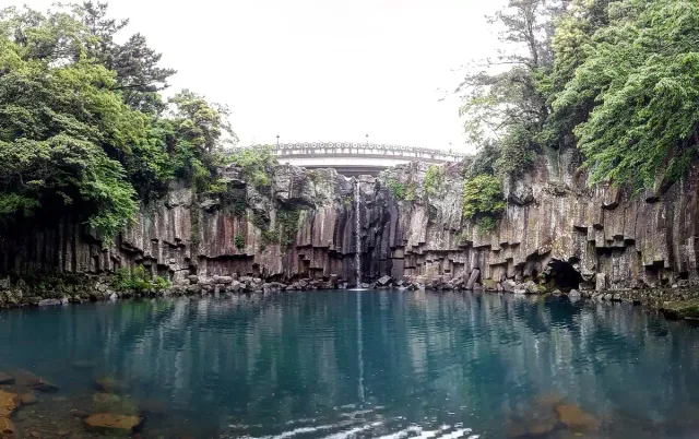 A turquoise pool at the foot of a columnar basalt cliff with an arched bridge above