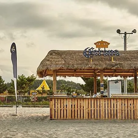 A wooden bar stand with a thatched roof on the beach sand, under a cloudy sky