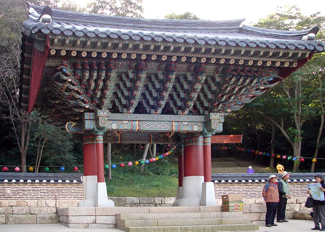 Temple gateway on red timber pillars with a heavy tiled roof and painted bracketing, paper lanterns strung across the yard