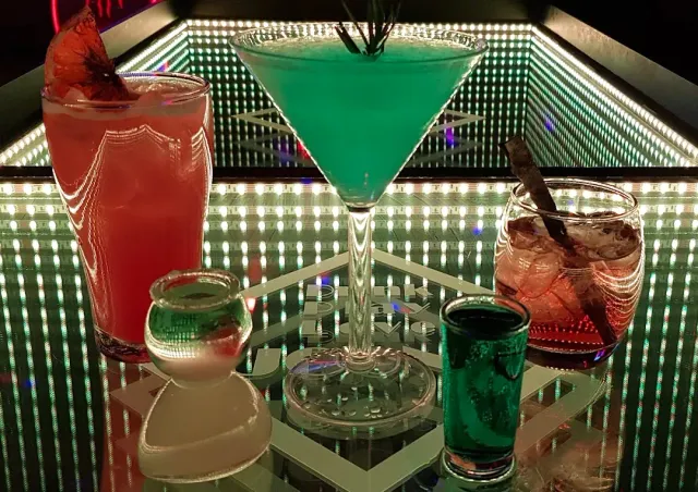 Four drinks on a mirrored table lit with bulbs, among them a green martini glass