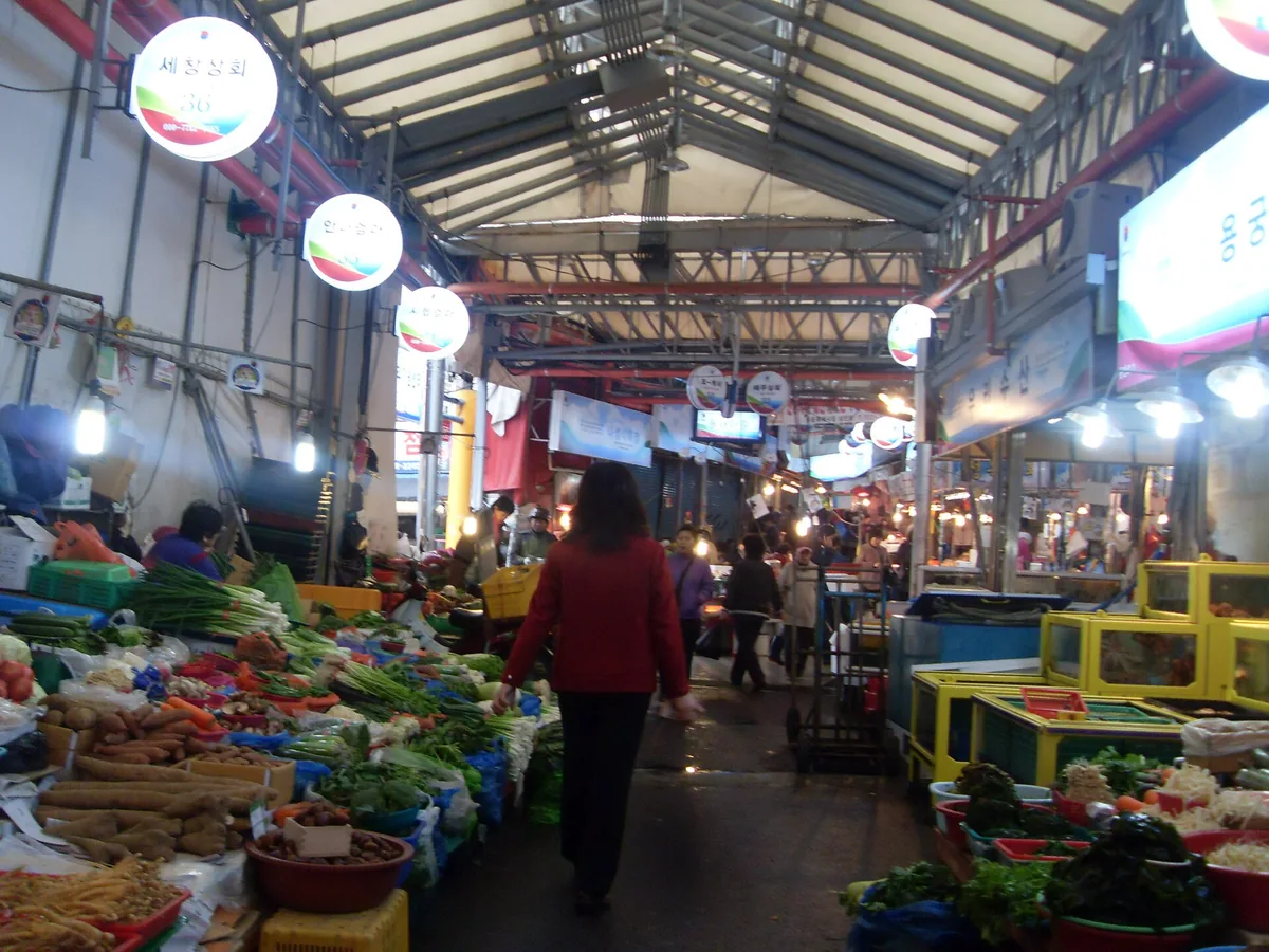 A covered market aisle with vegetable stalls on both sides and hanging lamps
