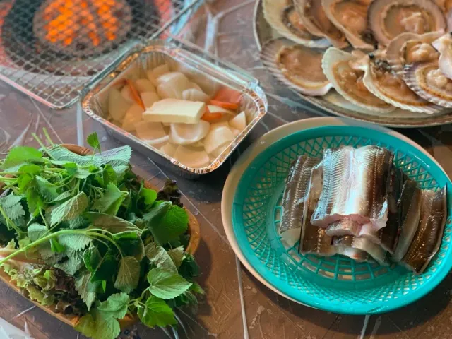 A grill table with scallops in their shells, fish fillets in a basket and a pile of green leaves