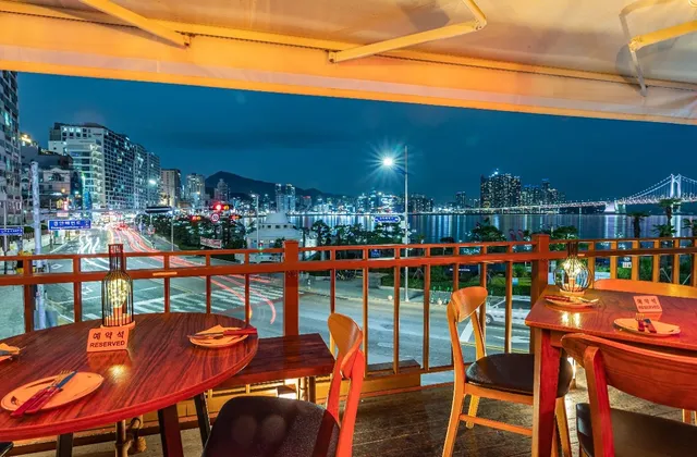 A balcony with wooden tables facing a bay, a skyline and a lit bridge at night