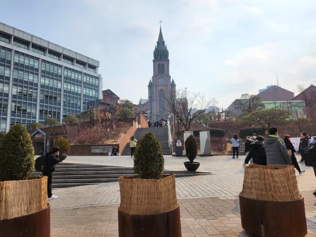 A brick church with a tall spire above a paved plaza and passers by