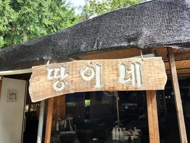 A carved wooden sign hanging under a shade net, with an open structure and trees behind it