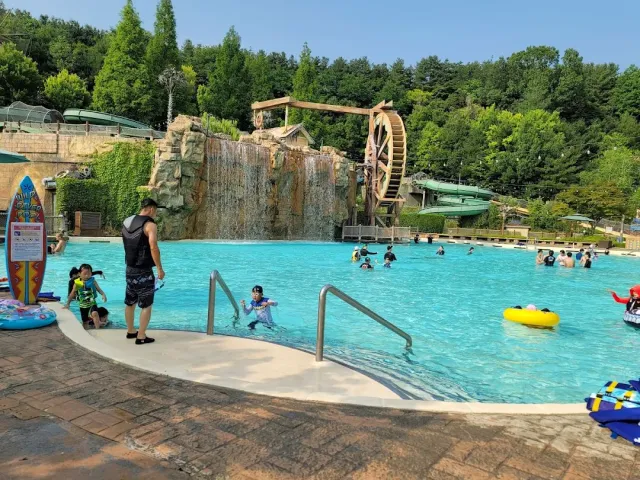 A turquoise pool with a rock waterfall and a water wheel, and bathers in the shallow water