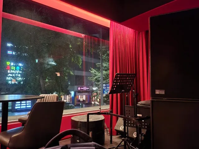 A room in red light with a window onto a street full of neon signs, a curtain and a music stand