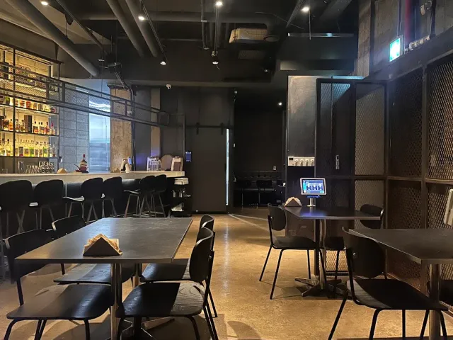 A dark industrial style space with black tables, stools along a counter and lit shelves