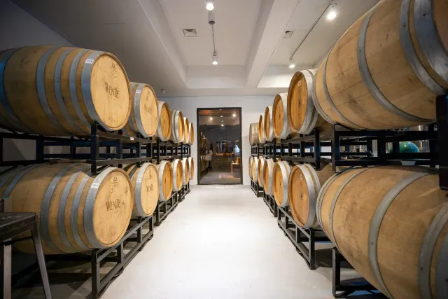 Two rows of wooden barrels on metal racks on both sides of a lit passage