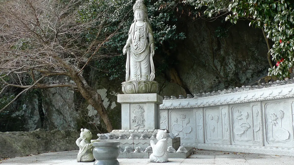 A stone statue of the goddess of mercy on a carved plinth, beside a stone balustrade and a wooded cliff